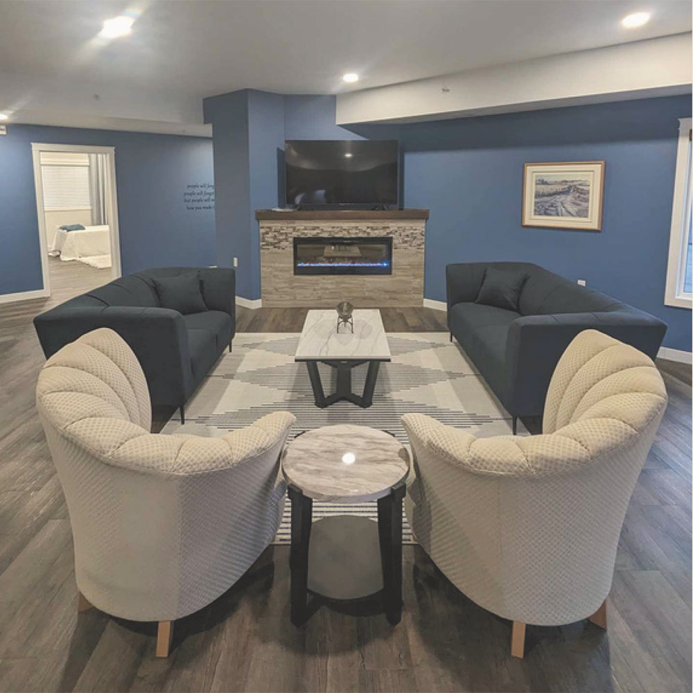 Common area of the Lyons Care assisted living housing facility. The walls are a rich blue, with laminate flooring in a pale wood. Comfy blue and beige chairs and loveseats and an end table surround a grey area rug and a contemporary coffee table with stone stop and black metal legs. In the background is a fireplace an large screen television.